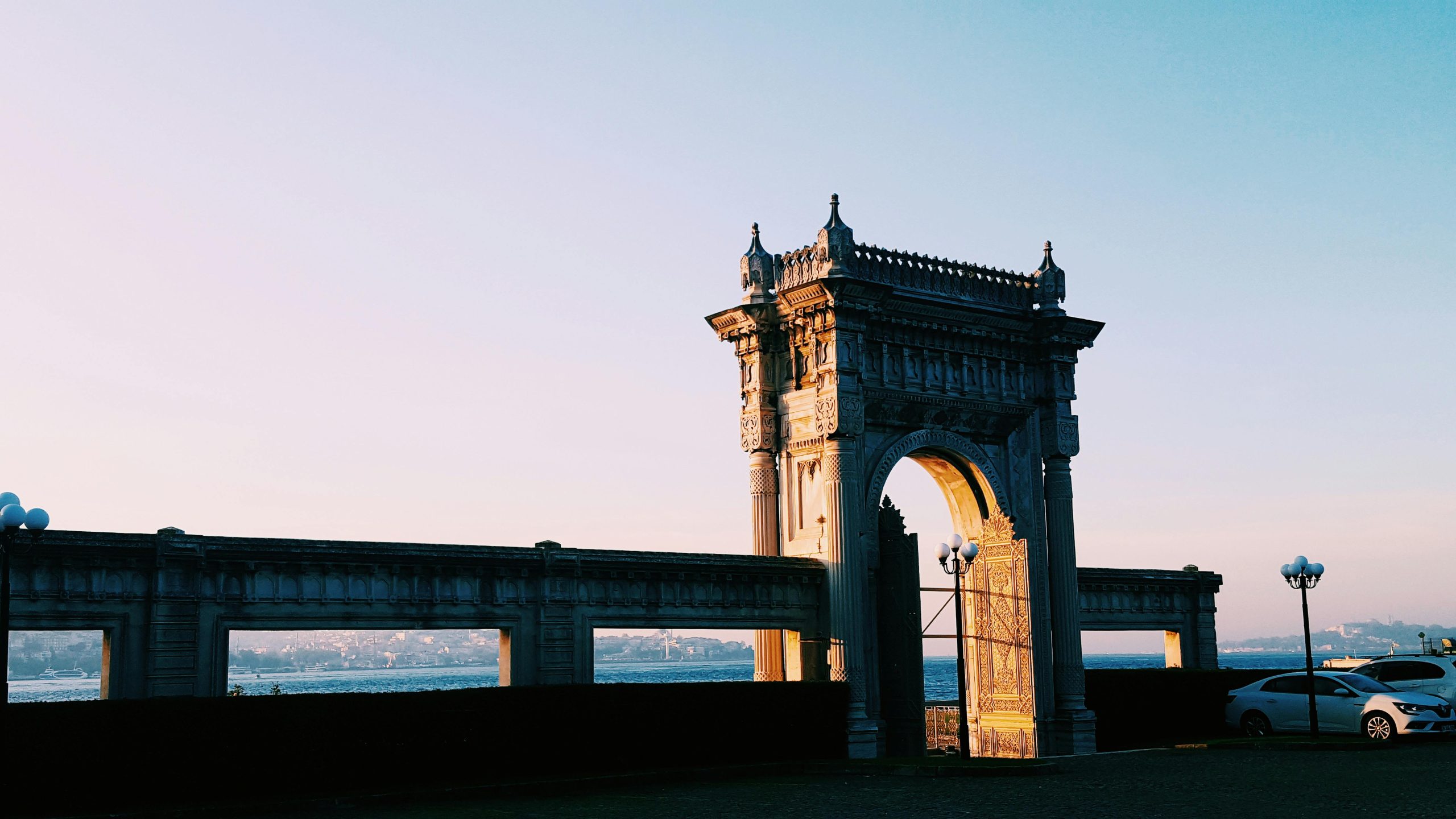 A beautifully lit historical gate by the Bosphorus in Istanbul during sunset with parked cars nearby.